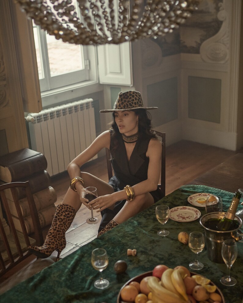 Op de afbeelding: Een vrouw met een hoed en laarzen met luipaardprint zit aan een tafel met een groen tafelkleed. Er staan glazen wijn, een fles champagne en een kom met fruit op tafel.