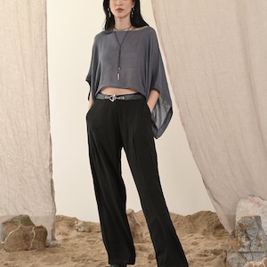 May include: A person wearing a grey, loose-fitting top and black wide-leg trousers. They are also wearing a black belt with a silver buckle and a long necklace. The background is a neutral colour with some rocks and sand.