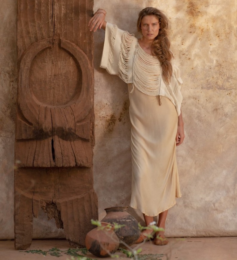 Pu&ograve; includere: Una donna indossa un top bianco lavorato a uncinetto con dettagli a frangia e una lunga gonna beige. &Egrave; in piedi accanto a una scultura in legno e tiene un vaso.