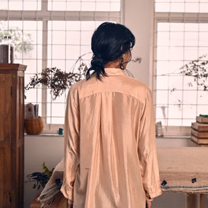 May include: A long-sleeved, peach-colored silk shirt with a collared neckline and button closure. The shirt is loose-fitting and has a relaxed silhouette.