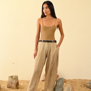 May include: A woman wearing a light brown tank top and beige linen pants with a brown leather belt. She is standing in a desert setting with rocks and sand.