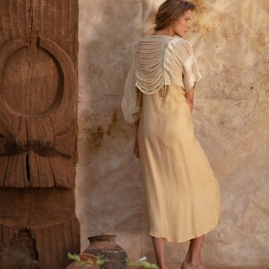 Pu&ograve; includere: Una donna indossa un abito color beige chiaro con un top in crochet bianco. Il top ha un design unico con la schiena scoperta e dettagli a frangia. La donna &egrave; in piedi davanti a un muro di legno con un design intagliato.