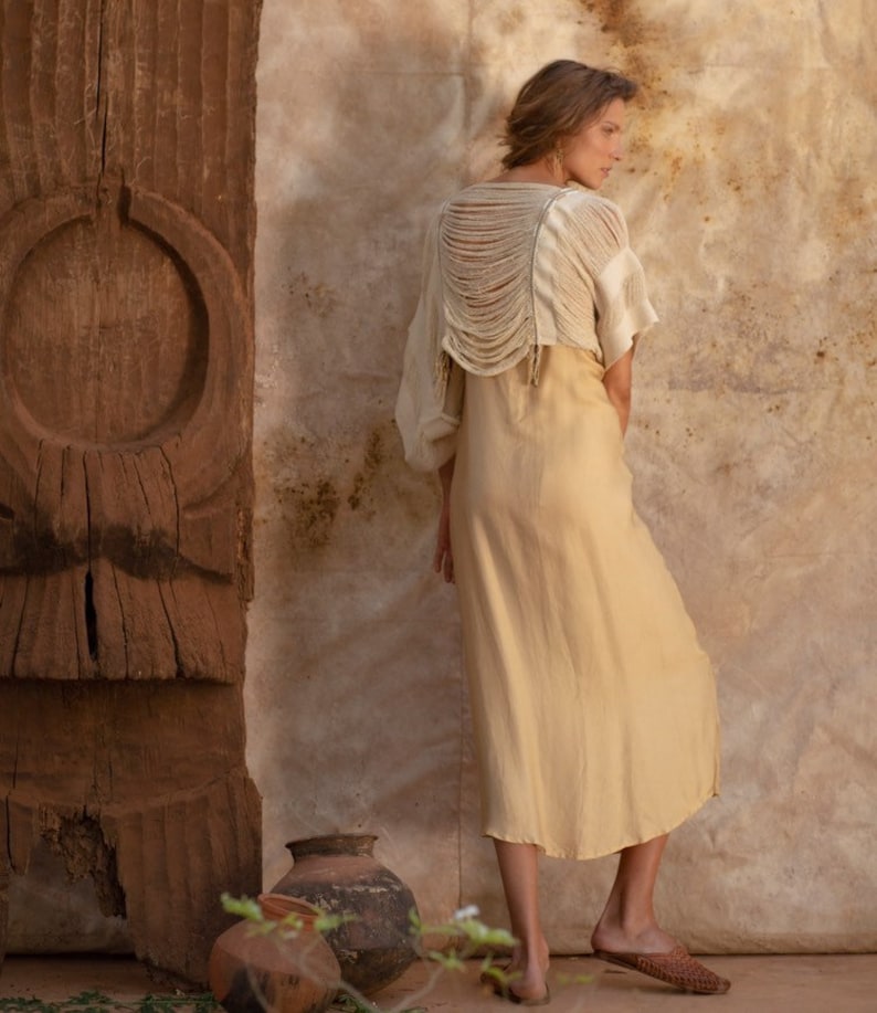 Pu&ograve; includere: Una donna indossa un abito color beige chiaro con un top in crochet bianco. Il top ha un design unico con la schiena scoperta e dettagli a frangia. La donna &egrave; in piedi davanti a un muro di legno con un design intagliato.