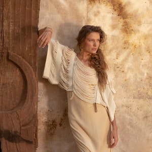 Pu&ograve; includere: Una donna indossa un top bianco all'uncinetto con dettagli a frange e una lunga gonna beige fluida. &Egrave; in piedi accanto a un muro di legno.