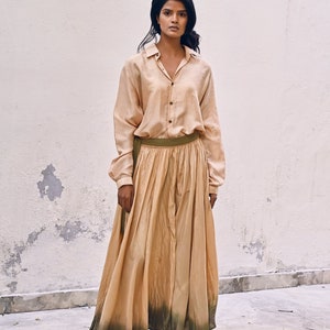 May include: A woman wearing a long, flowing, light brown skirt with a green ombre effect at the bottom. She is also wearing a light brown button-down shirt and brown shoes.