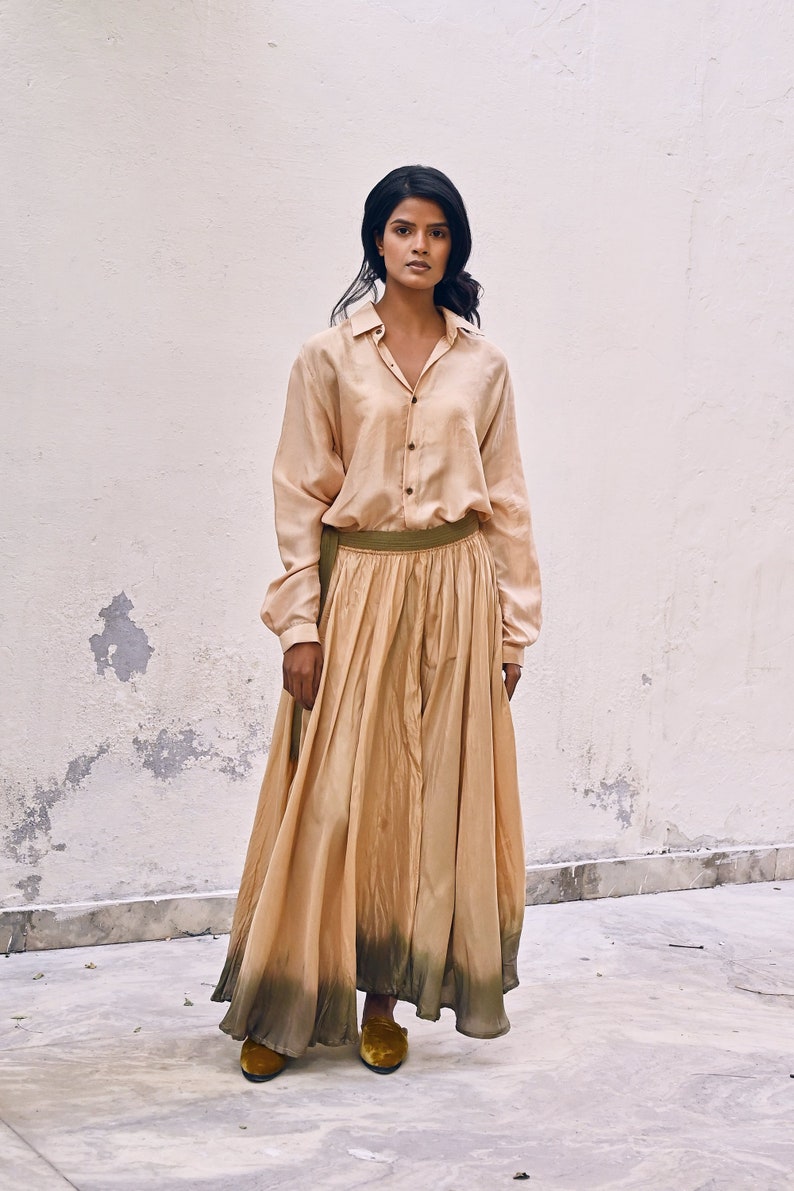 May include: A woman wearing a long, flowing, light brown skirt with a green ombre effect at the bottom. She is also wearing a light brown button-down shirt and brown shoes.