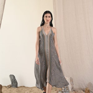 May include: A woman wearing a gray and blue patterned midi dress with a button-up front and a slit in the skirt. She is walking on a sandy surface with rocks in the background.