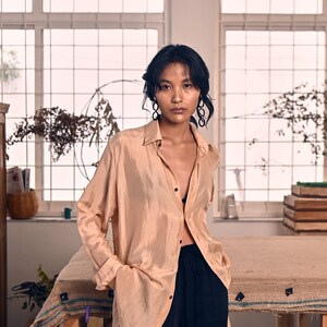 May include: A woman wearing a light brown, long-sleeved shirt with a black bra peeking out from the neckline. She is wearing black pants and has her hair pulled back. She is standing in front of a window with a wooden table in front of her.