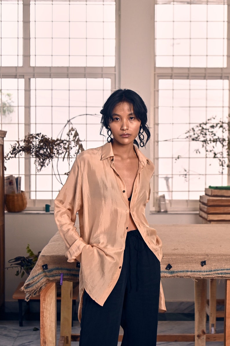 May include: A woman wearing a light brown, long-sleeved shirt with a black bra peeking out from the neckline. She is wearing black pants and has her hair pulled back. She is standing in front of a window with a wooden table in front of her.
