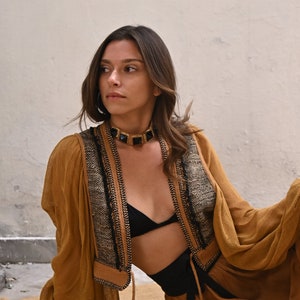 May include: A woman wearing a brown, long-sleeved top with black trim and a black bra. She is wearing a gold and black choker necklace and a black belt.