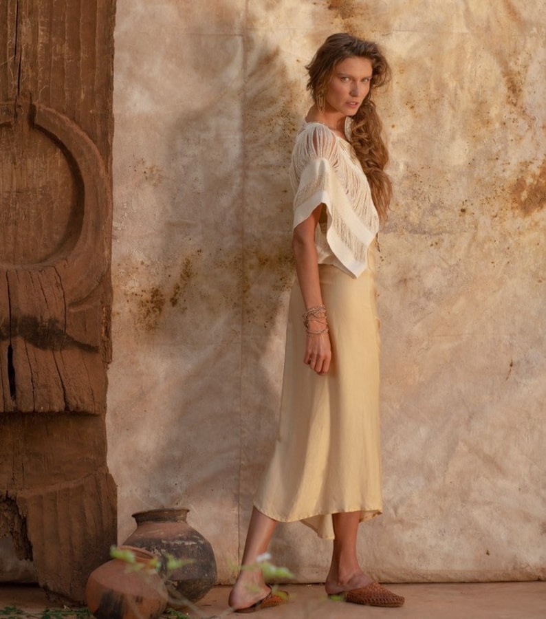 Pu&ograve; includere: Una donna indossa un top bianco lavorato a uncinetto e una gonna beige, in piedi davanti a un muro di legno rustico. Indossa sandali marroni e ha lunghi capelli castani.