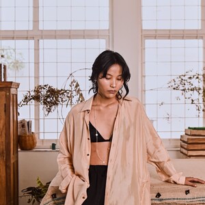 May include: A woman wearing a light peach colored shirt over a black bra and black pants. She is wearing a gold necklace with a pendant. The shirt is open at the front and the woman is standing in front of a window.