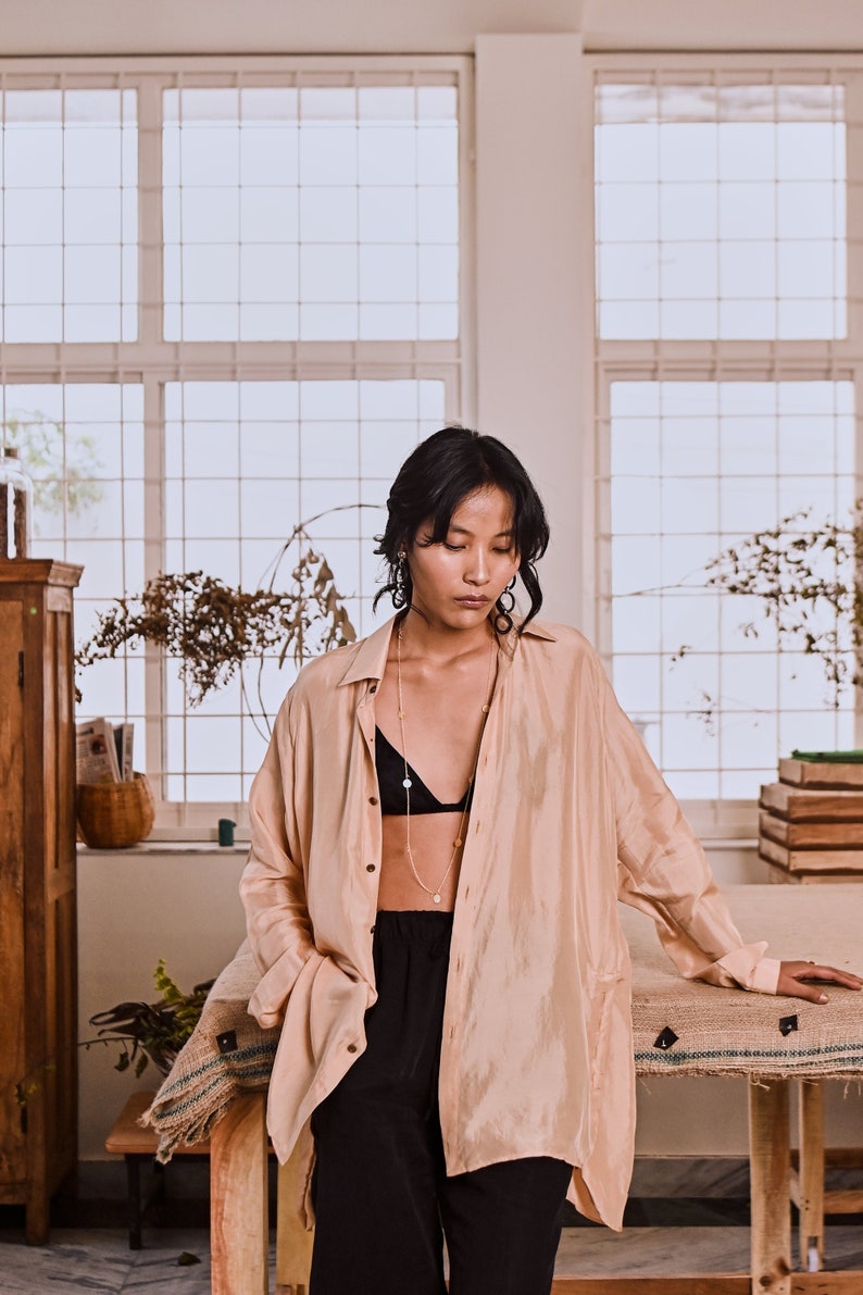 May include: A woman wearing a light peach colored shirt over a black bra and black pants. She is wearing a gold necklace with a pendant. The shirt is open at the front and the woman is standing in front of a window.