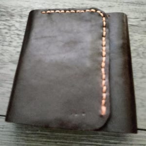 May include: A dark brown leather wallet with a curved flap and visible tan stitching along the edges. The wallet appears to be folded, with a simple, minimalist design. The leather has a slightly textured appearance.