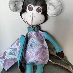 May include: Handmade monkey doll with a black head, white face, and large, round ears. The doll wears a teal collar and a patterned dress in shades of purple and gray. Teal arms and legs complete the whimsical design. A brown strap is attached to the doll.