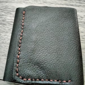 May include: A dark grey leather wallet with a textured surface. The edges are stitched with a contrasting pink thread. The wallet is folded in half, suggesting a compact design for carrying essentials.