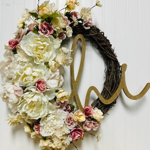 May include: A floral wreath with a woven vine base, decorated with cream, pink, and burgundy artificial flowers. A wooden "love" script is attached to the wreath, creating a decorative piece.