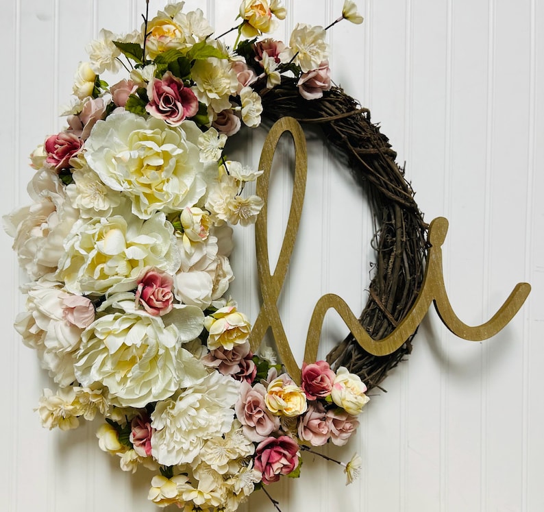 May include: A floral wreath with a woven vine base, decorated with cream, pink, and burgundy artificial flowers. A wooden "love" script is attached to the wreath, creating a decorative piece.
