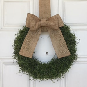 May include: A round, green moss wreath with a large burlap bow and hanging ribbon. The wreath is displayed on a white door. The burlap bow and ribbon add a rustic touch to the natural green wreath, suitable for home decor.