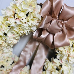 Peut inclure: Une couronne florale orn&eacute;e de fleurs d'hortensia cr&egrave;me avec de subtils accents roses. Un grand n&oelig;ud en ruban de satin de couleur blush ajoute une touche d'&eacute;l&eacute;gance. Cette couronne d&eacute;corative est parfaite pour ajouter une touche printani&egrave;re &agrave; n'importe quelle maison.