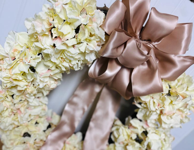 Peut inclure: Une couronne florale orn&eacute;e de fleurs d'hortensia cr&egrave;me avec de subtils accents roses. Un grand n&oelig;ud en ruban de satin de couleur blush ajoute une touche d'&eacute;l&eacute;gance. Cette couronne d&eacute;corative est parfaite pour ajouter une touche printani&egrave;re &agrave; n'importe quelle maison.