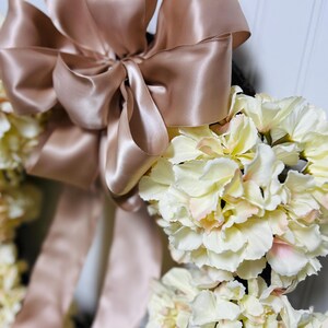 Peut inclure: Une couronne florale orn&eacute;e de fleurs cr&egrave;me avec de l&eacute;gers accents roses. Un grand n&oelig;ud en satin rose dor&eacute; est plac&eacute; en haut. Cet &eacute;l&eacute;ment d&eacute;coratif est id&eacute;al pour les c&eacute;l&eacute;brations et les d&eacute;corations int&eacute;rieures.
