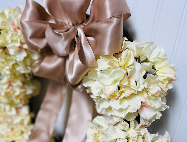 Peut inclure: Une couronne florale orn&eacute;e de fleurs cr&egrave;me avec de l&eacute;gers accents roses. Un grand n&oelig;ud en satin rose dor&eacute; est plac&eacute; en haut. Cet &eacute;l&eacute;ment d&eacute;coratif est id&eacute;al pour les c&eacute;l&eacute;brations et les d&eacute;corations int&eacute;rieures.