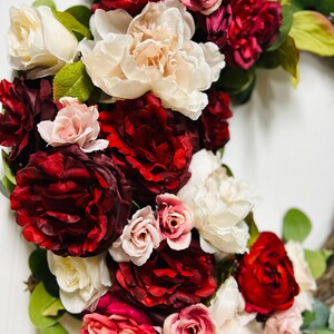以下が含まれることがあります： 赤、ピンク、クリーム色の花が咲き誇る花のリース。バラやシャクヤクを含み、緑の葉が散りばめられています。花は密集しており、豊かで鮮やかなディスプレイを作り出しています。装飾品です。