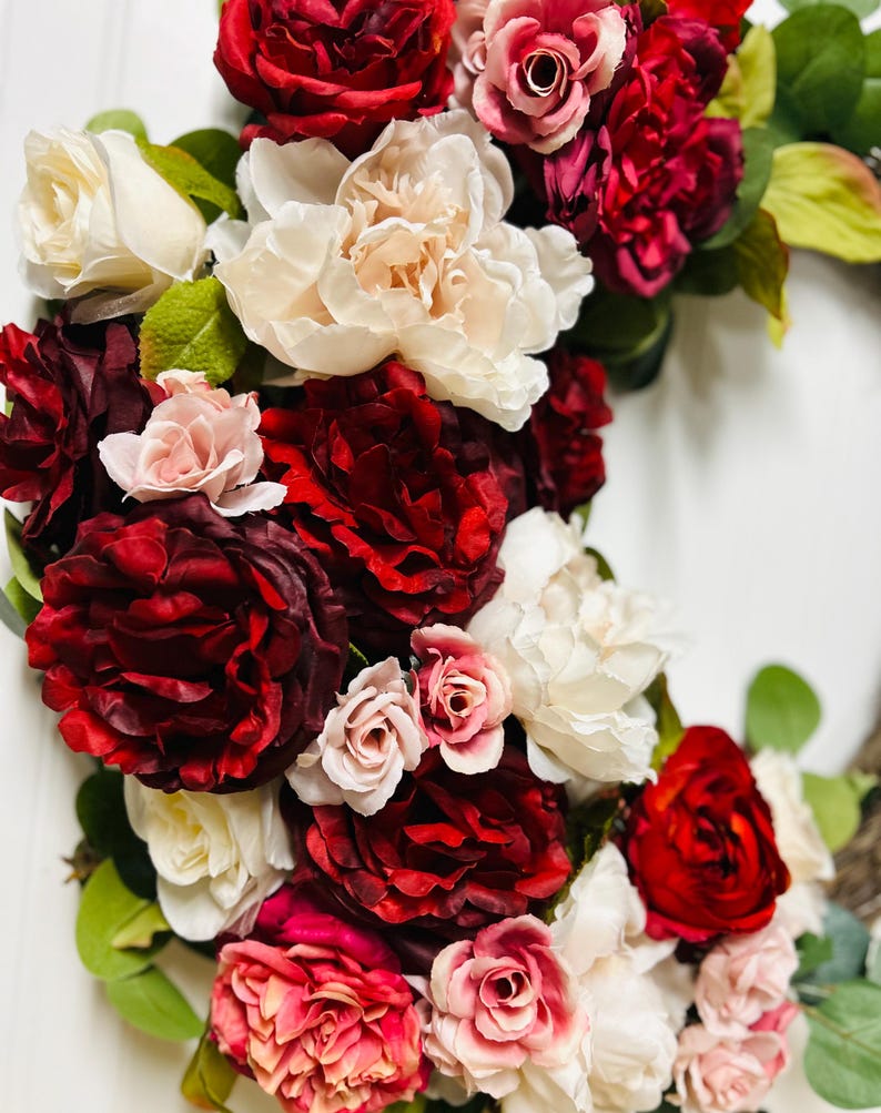 以下が含まれることがあります： 赤、ピンク、クリーム色の花が咲き誇る花のリース。バラやシャクヤクを含み、緑の葉が散りばめられています。花は密集しており、豊かで鮮やかなディスプレイを作り出しています。装飾品です。