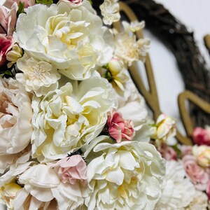 May include: A close-up of a floral wreath with large, creamy white peonies and smaller pink and red roses. The wreath is set against a white background, with a brown vine base and gold-coloured letters.