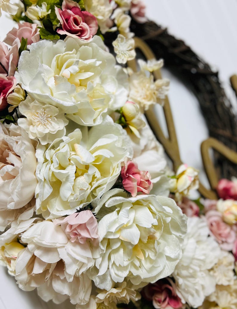 May include: A close-up of a floral wreath with large, creamy white peonies and smaller pink and red roses. The wreath is set against a white background, with a brown vine base and gold-coloured letters.