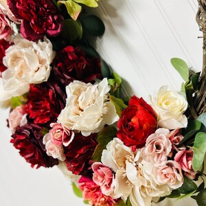 以下が含まれることがあります： 赤、ピンク、クリーム色の人工花を使用した花のリース。 リースは編まれた小枝で作られ、緑の葉が含まれています。 鮮やかで装飾的なアクセントで、直径約30cmです。