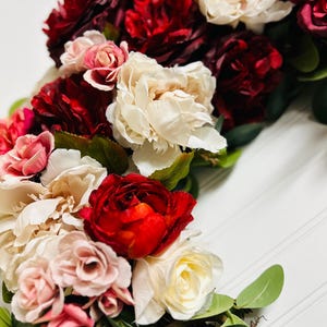 以下が含まれることがあります： 赤、ピンク、クリーム色の花が咲き誇る花のリースをクローズアップ。 リースは緑の葉で飾られており、自然素材で作られているように見え、素朴でエレガントな美しさを醸し出しています。