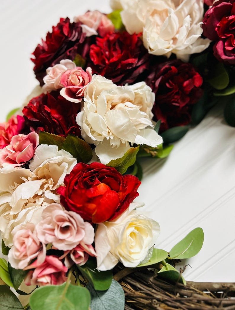 以下が含まれることがあります： 赤、ピンク、クリーム色の花が咲き誇る花のリースをクローズアップ。 リースは緑の葉で飾られており、自然素材で作られているように見え、素朴でエレガントな美しさを醸し出しています。