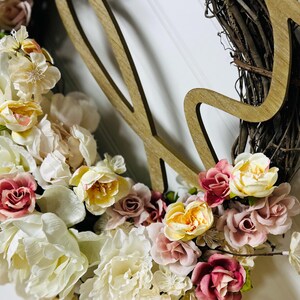 May include: A floral wreath with a wooden accent. The wreath is made of woven twigs and adorned with cream, yellow, and pink flowers. A wooden cutout of the word "Love" is attached to the wreath.