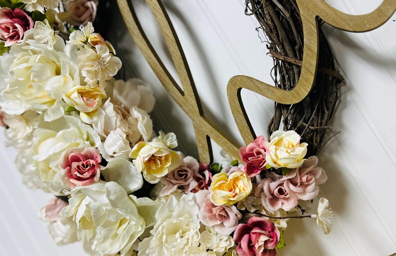 May include: A floral wreath with a wooden accent. The wreath is made of woven twigs and adorned with cream, yellow, and pink flowers. A wooden cutout of the word "Love" is attached to the wreath.