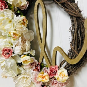 May include: A decorative wreath featuring a wooden 'H' and a twig base. The wreath is decorated with artificial flowers in white, cream, pink, and yellow, creating a soft, inviting aesthetic. The wreath is suitable for indoor decoration.