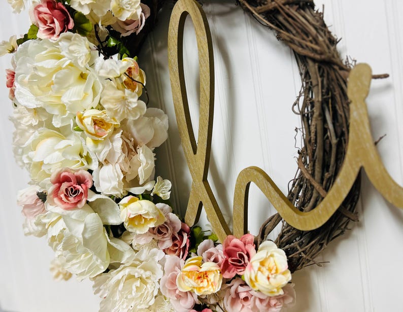 May include: A decorative wreath featuring a wooden 'H' and a twig base. The wreath is decorated with artificial flowers in white, cream, pink, and yellow, creating a soft, inviting aesthetic. The wreath is suitable for indoor decoration.