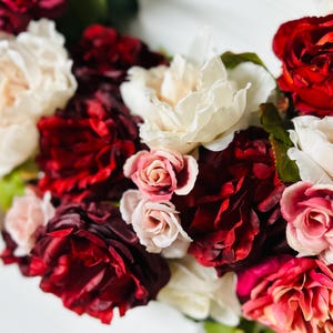 以下が含まれることがあります： 赤、白、ピンクの色合いの花々が咲き誇る、花の配置のクローズアップ。バラやその他の花々、緑の葉が散りばめられています。