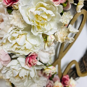 May include: A floral wreath with large, creamy white and pink peonies and smaller accent flowers. The wreath has a grapevine base and a wooden cutout spelling "love". The flowers are densely clustered, creating a full, textured appearance.