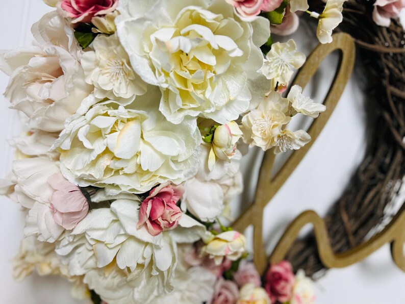 May include: A floral wreath with large, creamy white and pink peonies and smaller accent flowers. The wreath has a grapevine base and a wooden cutout spelling "love". The flowers are densely clustered, creating a full, textured appearance.