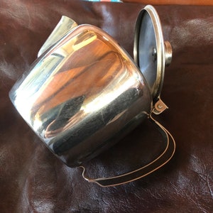 May include: A silver stainless steel teapot with a hinged lid and a black handle. The teapot is sitting on a brown leather surface.