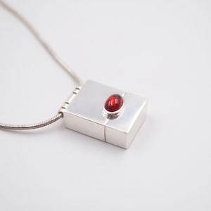May include: A silver necklace with a small rectangular locket pendant. The locket has a red gemstone set into the top.
