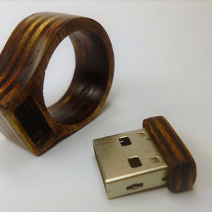 Op de afbeelding: Een houten ring met een USB-stick eraan bevestigd. De ring is gemaakt van donker hout met lichtbruine strepen. De USB-stick is zilverkleurig met een donkerhouten hoes.