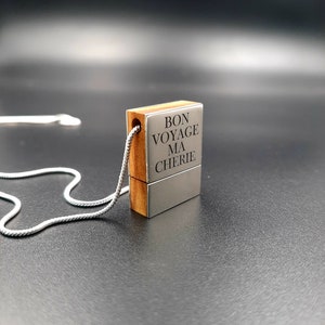 Op de afbeelding: Een rechthoekige hangerketting met een zilveren ketting. De hanger combineert hout en zilverkleurig metaal, met de Franse zin "BON VOYAGE MA CHERIE" gegraveerd op het metaal. De ketting wordt getoond tegen een donkere achtergrond.