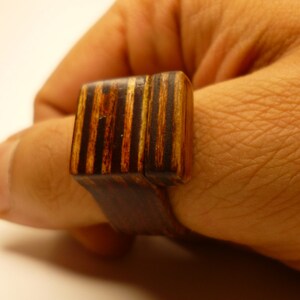 Op de afbeelding: Een houten ring met een vierkante bovenkant gemaakt van gelaagd hout met bruine en zwarte strepen. De ring wordt aan een vinger gedragen.