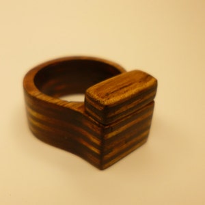 Op de afbeelding: Een houten ring met een uniek ontwerp. De ring is gemaakt van donker hout met lichtbruine strepen. De ring heeft een vierkant stuk hout dat aan de zijkant is bevestigd.