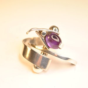 Könnte beinhalten: Ein silberner Ring mit einem violetten Amethyst-Edelstein, der in einem geometrischen Design gefasst ist. Der Ring hat ein verdrehtes Band und zwei silberne Kugeln.