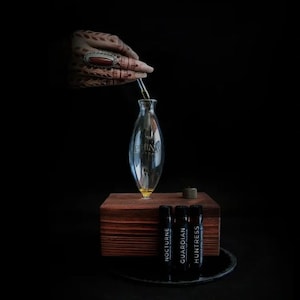 Peut inclure: Une bouteille diffuseur en verre transparent avec une base en bois et trois petites bouteilles noires étiquetées "Nocturne", "Guardian" et "Huntress".