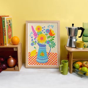 May include: A framed print of a colorful floral arrangement in a blue pitcher with a gingham tablecloth background. The flowers are pink, yellow, orange, and red. The pitcher has a blue and white pattern.